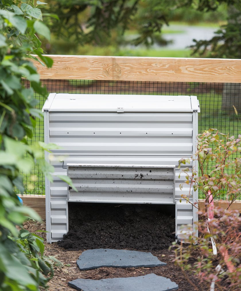 Demeter Metal Compost Bin in grey in a garden with bottom flap open and soil inside